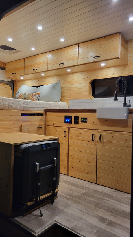 This cozy RV rental interior features a wood finish, kitchen with sink, bed, and pull-out fridge, perfect for van life adventures.