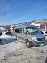 Silver RV rental van with 'SKI HUT' plate, parked in a snowy mountain resort on a sunny winter day.