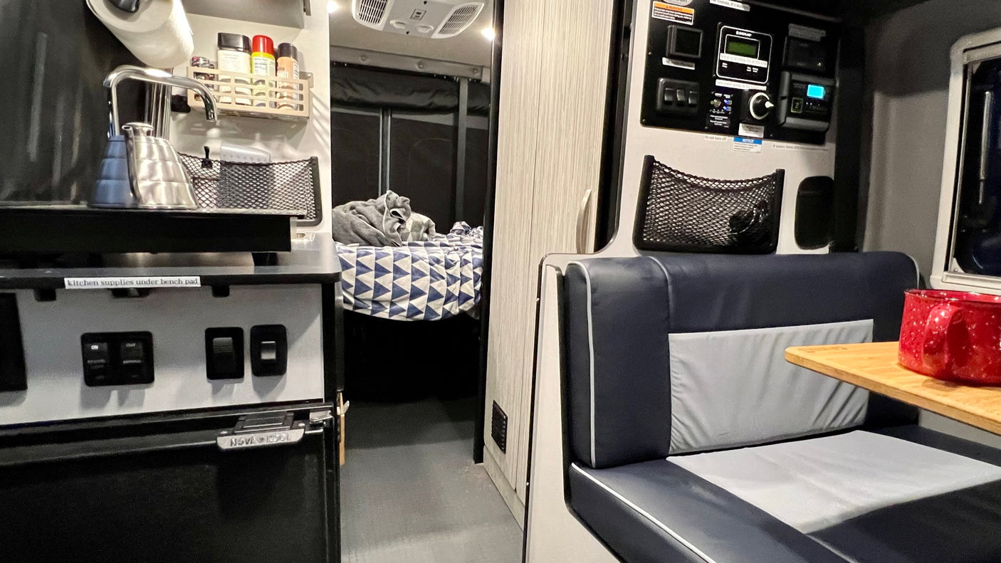 Inside a compact RV rental, featuring a kitchen area with kettle, a bed, and a small dining bench with a red mug.