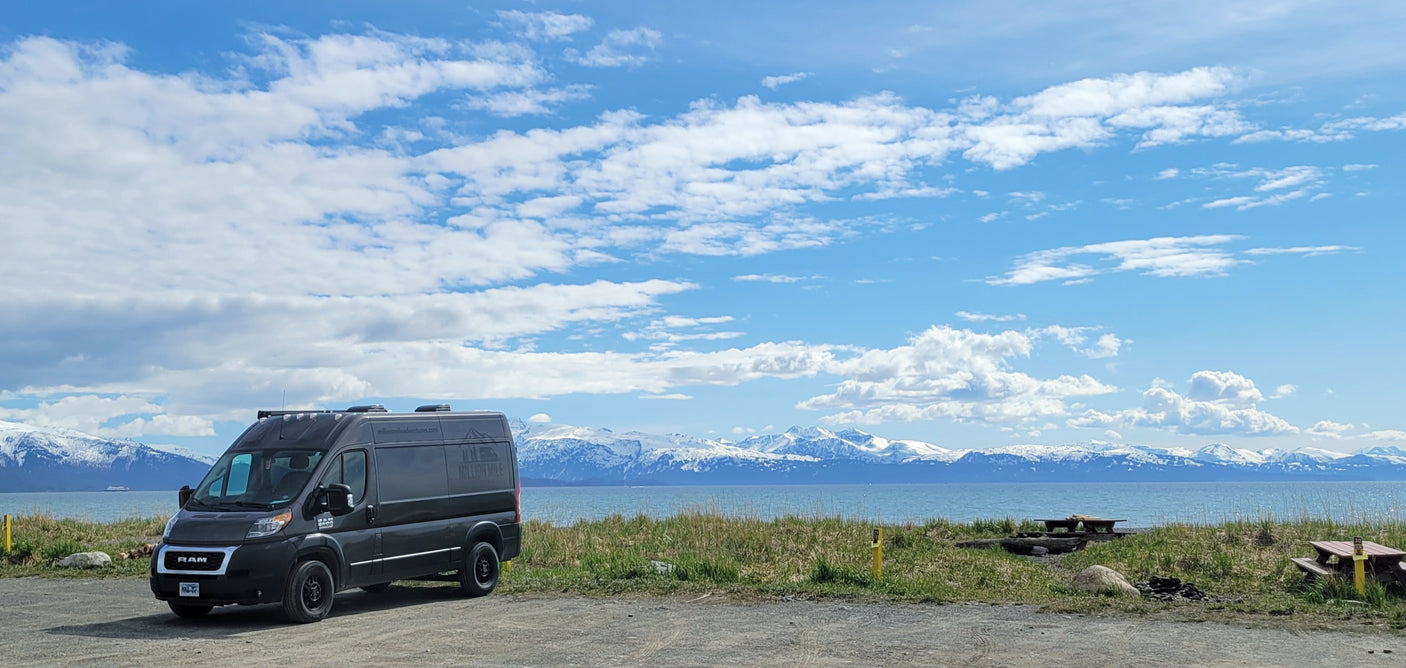 Dark grey RV rental van at a scenic waterfront campsite, with snow-capped mountains, a distant ship, and bright blue sky.