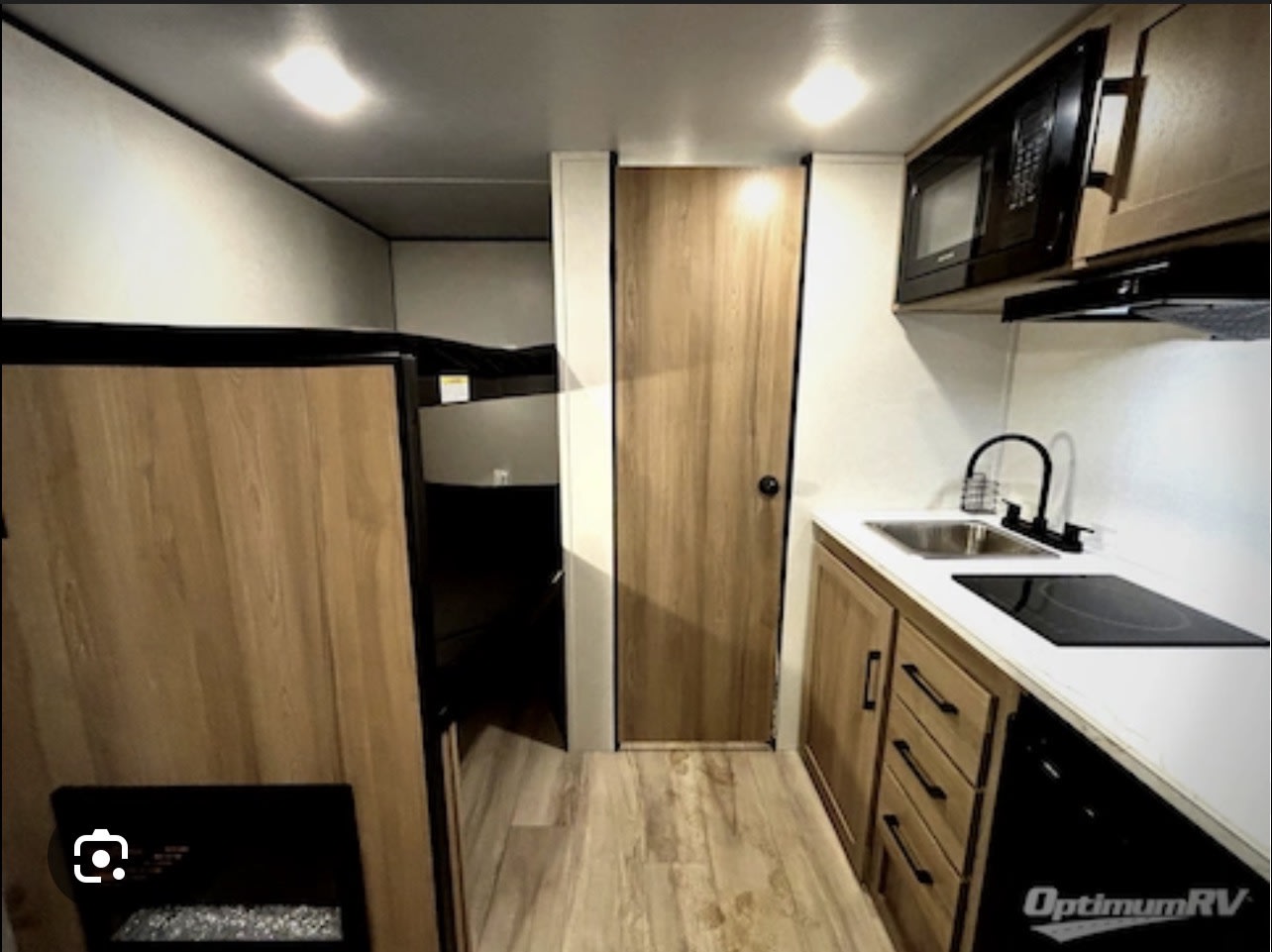 RV rental interior with a compact kitchenette featuring a sink, stovetop, microwave, and light wood cabinets. A door and bunk beds are visible.