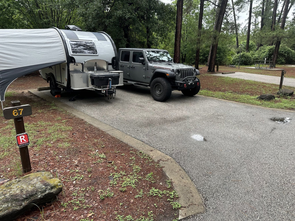  with vehicles parked among tall trees.|This RV rental features a gray Jeep Gladiator towing a teardrop camper at campsite 67 — photo 7