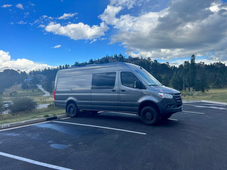 A sleek gray Mercedes-Benz Sprinter van, perfect for an RV rental, parked in a scenic mountain lot under a partly cloudy sky.