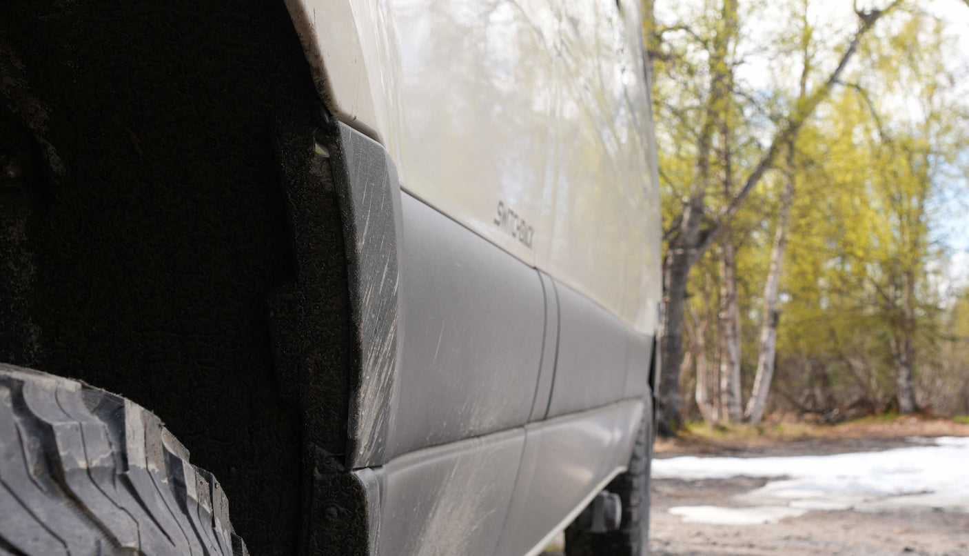 Close-up of a muddy RV rental's side, showing its tire, wheel well, and dirty panels, set in a snowy, wooded area.