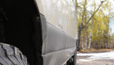 Close-up of a muddy RV rental's side, showing its tire, wheel well, and dirty panels, set in a snowy, wooded area.