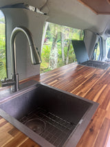 Inside an RV rental: a modern van kitchen with a stainless steel sink, wood countertop, and scenic tropical view.