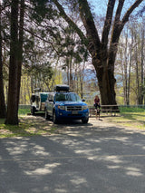 Blue Ford Escape towing a teardrop trailer (RV rental) at a shaded campsite. A woman and dog relax by a picnic table amidst trees.