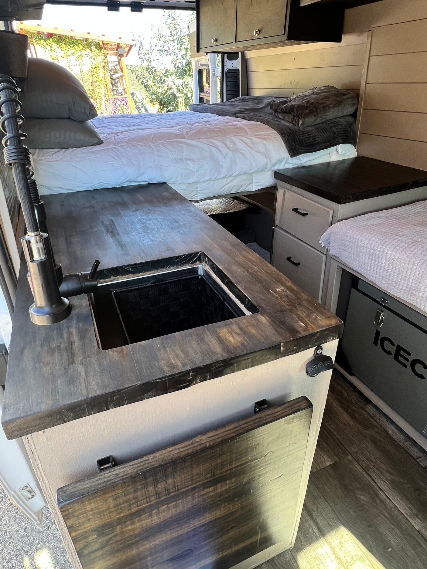 Inside a stylish RV rental, featuring a cozy bed, dark wood kitchenette with sink, and an ICECO cooler.