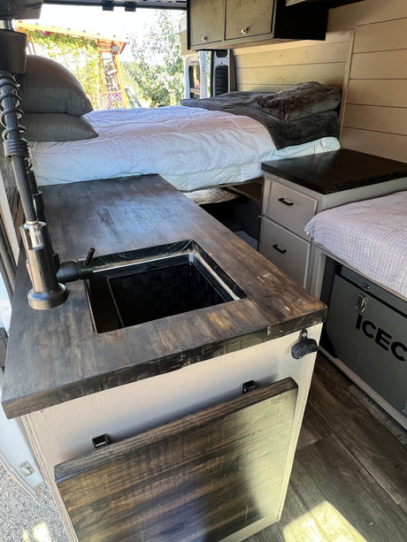 Inside a stylish RV rental, featuring a cozy bed, dark wood kitchenette with sink, and an ICECO cooler.