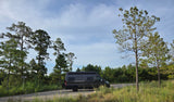 A dark grey rv rental parked by tall pine trees on a roadside, surrounded by green foliage under a blue sky.