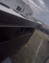 Side view of a black vessel/trailer parked, with a white RV rental behind it on a gravel lot under a cloudy sky.