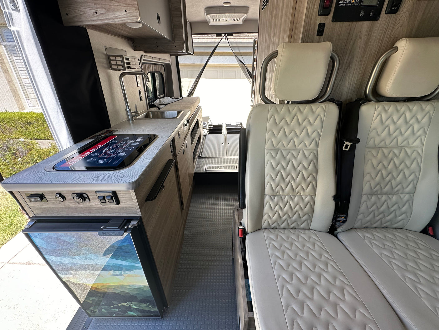 Modern rv rental interior: compact kitchen with sink, stovetop & fridge; comfortable passenger seats. Rear door open to outside.