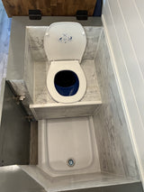 Compact RV rental bathroom: white toilet with blue bucket, white shower pan, and marbled gray walls in a wet bath setup.