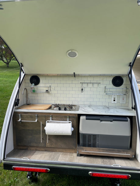 A well-equipped outdoor kitchen in an rv rental teardrop camper, showing a sink, stove, storage, and portable fridge with a marble countertop.