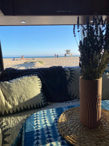 Cozy RV rental interior with pillows, blanket, and lavender, overlooking a sunny beach with ocean, sand, and lifeguard tower.