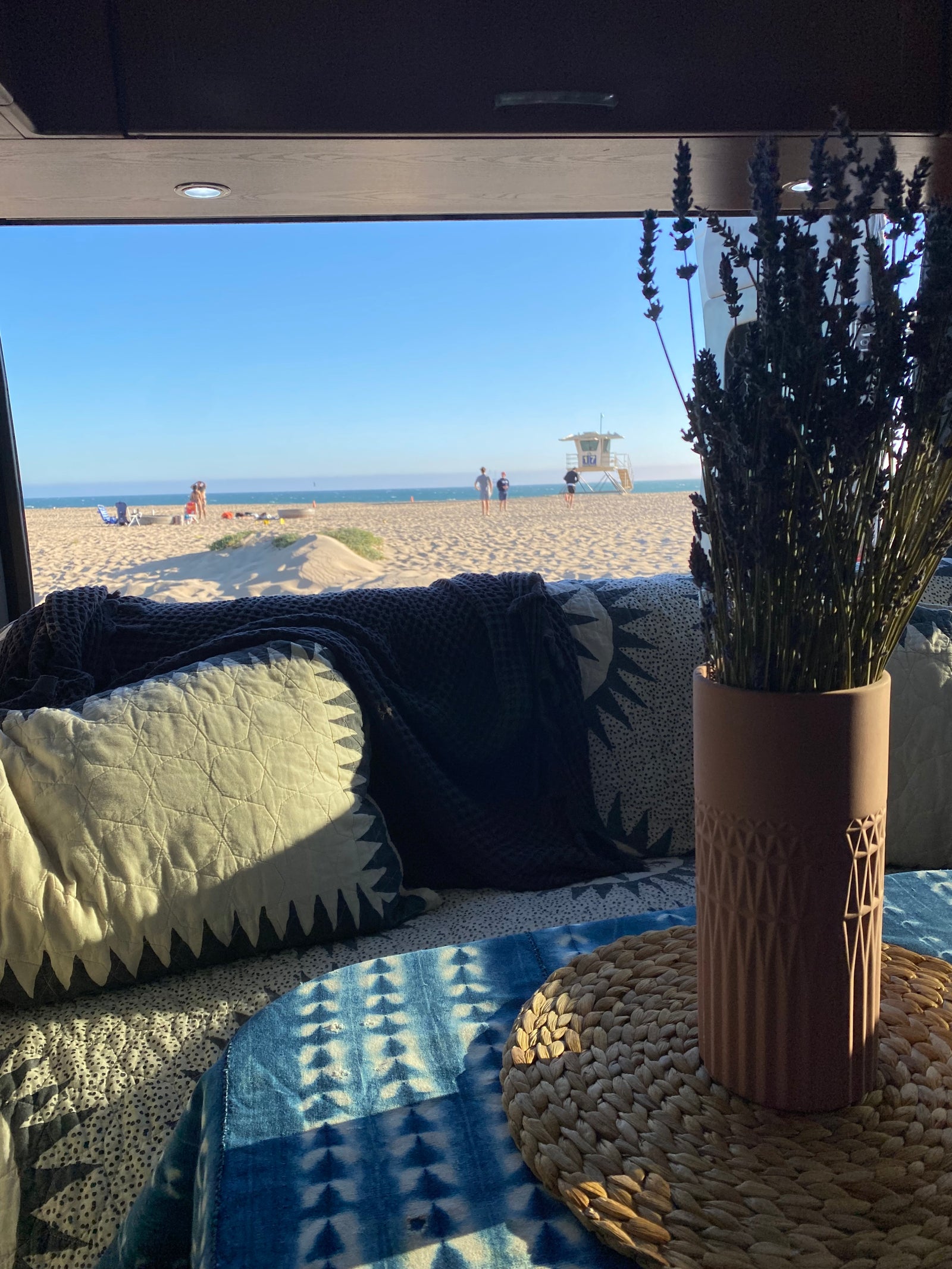 Cozy RV rental interior with pillows, blanket, and lavender, overlooking a sunny beach with ocean, sand, and lifeguard tower.