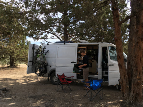 This rv rental offers adventure! Man cooks by white camper van with bikes on back, chairs set for outdoor living.