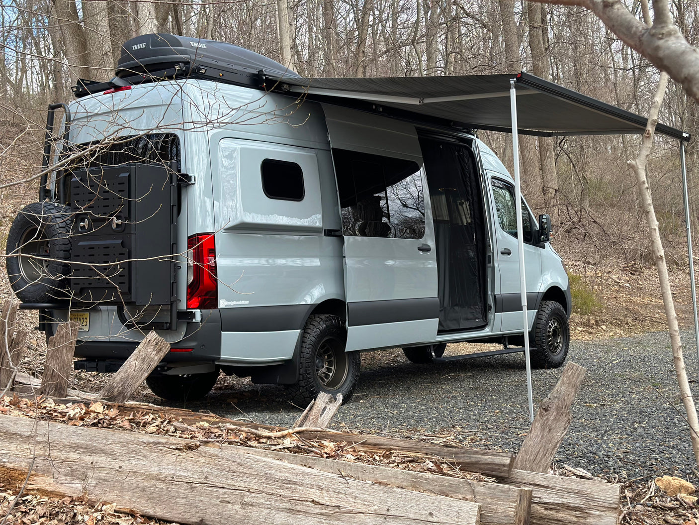 Overland-ready grey camper van with awning extended, perfect for an RV rental adventure in the woods. Enjoy nature!