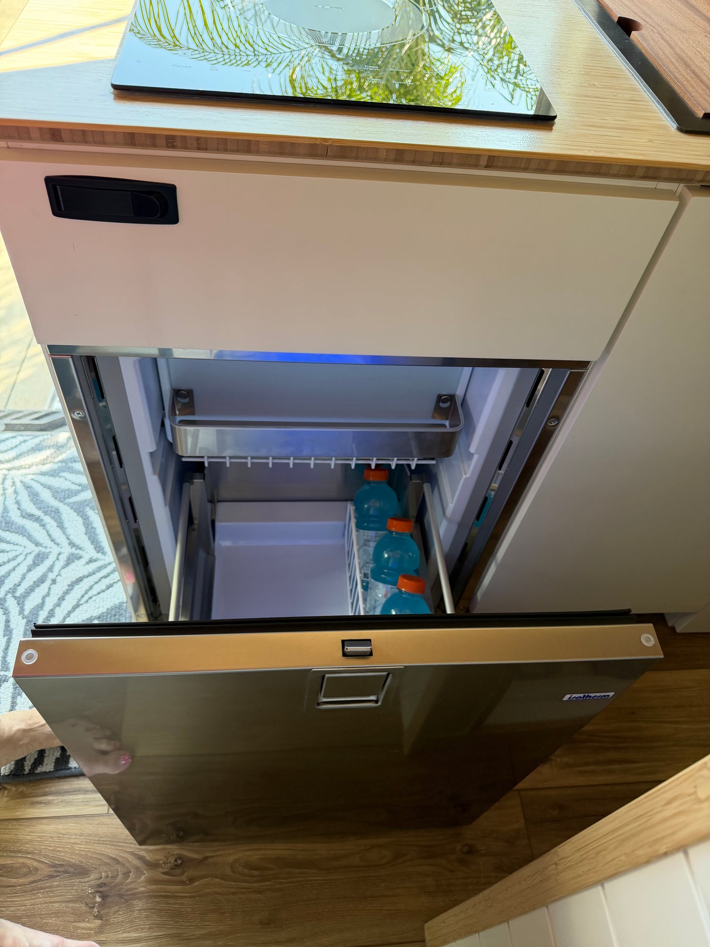 An open built-in drawer fridge in an RV rental kitchen, revealing blue Gatorade bottles. Above, a sleek cooktop reflects palm trees.