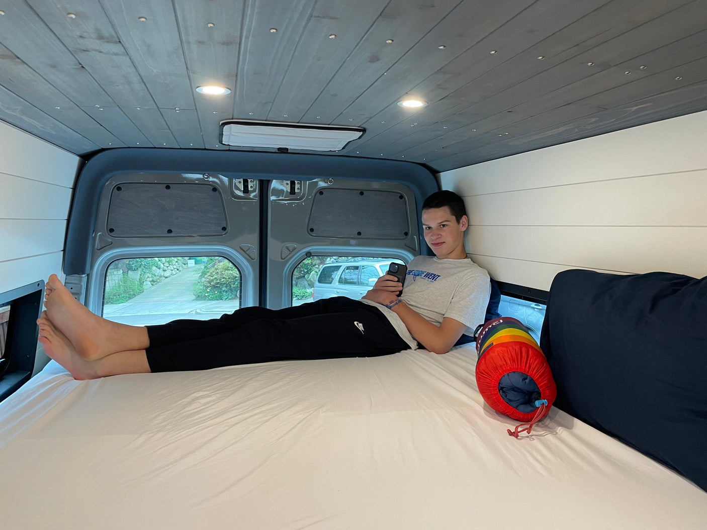 Young man enjoying comfort in an RV rental. He's lying on a white bed in a custom van, holding his phone. Cozy interior with wood ceiling.