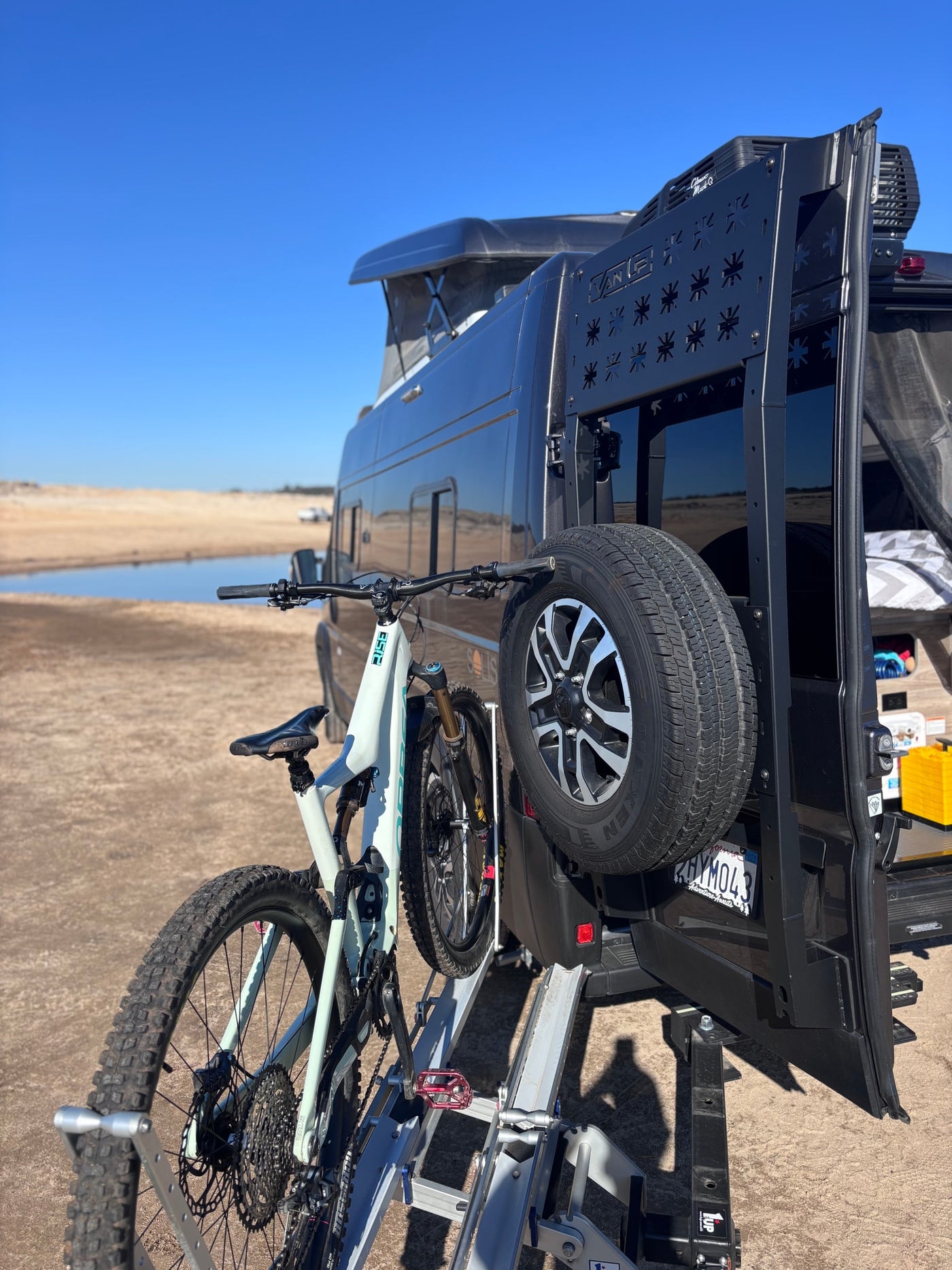 Adventure awaits! This RV rental features a pop-top van, bike rack with mountain bike, and spare tire, parked by water. — photo 9