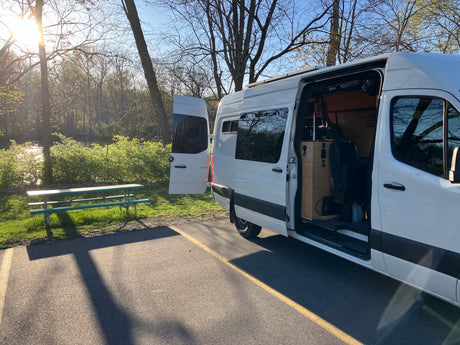 A white camper van, perfect for your next RV rental, is parked near a tranquil river and picnic table under sunlight.