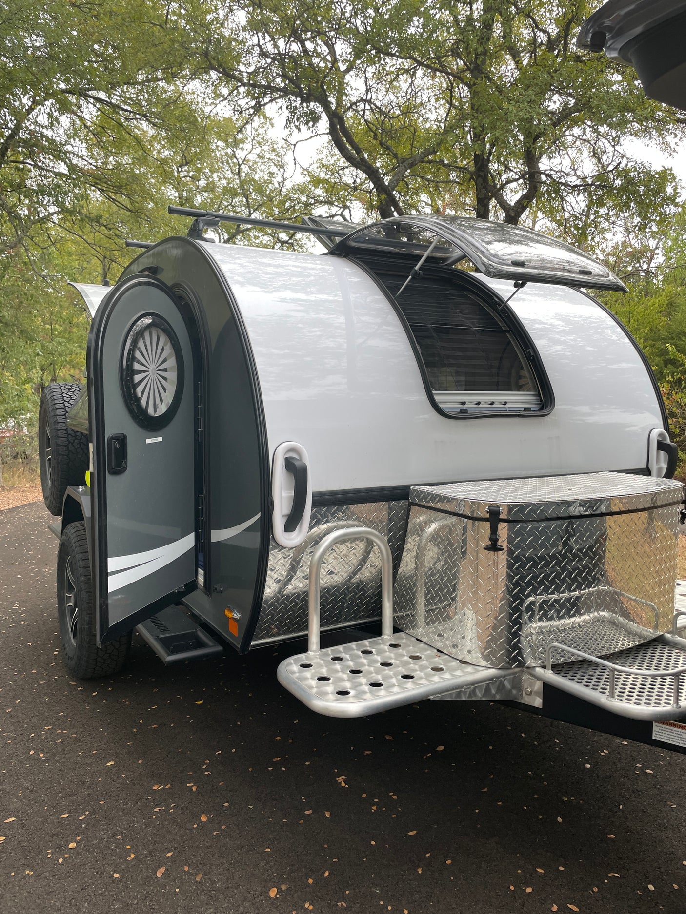 A stylish silver and gray teardrop camper RV rental, with its door and window open, features a diamond-plate storage box and steps.