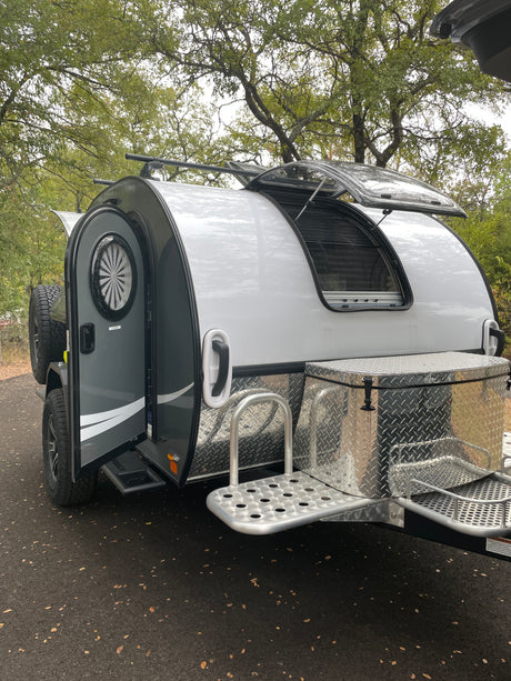 A stylish silver and gray teardrop camper RV rental, with its door and window open, features a diamond-plate storage box and steps.