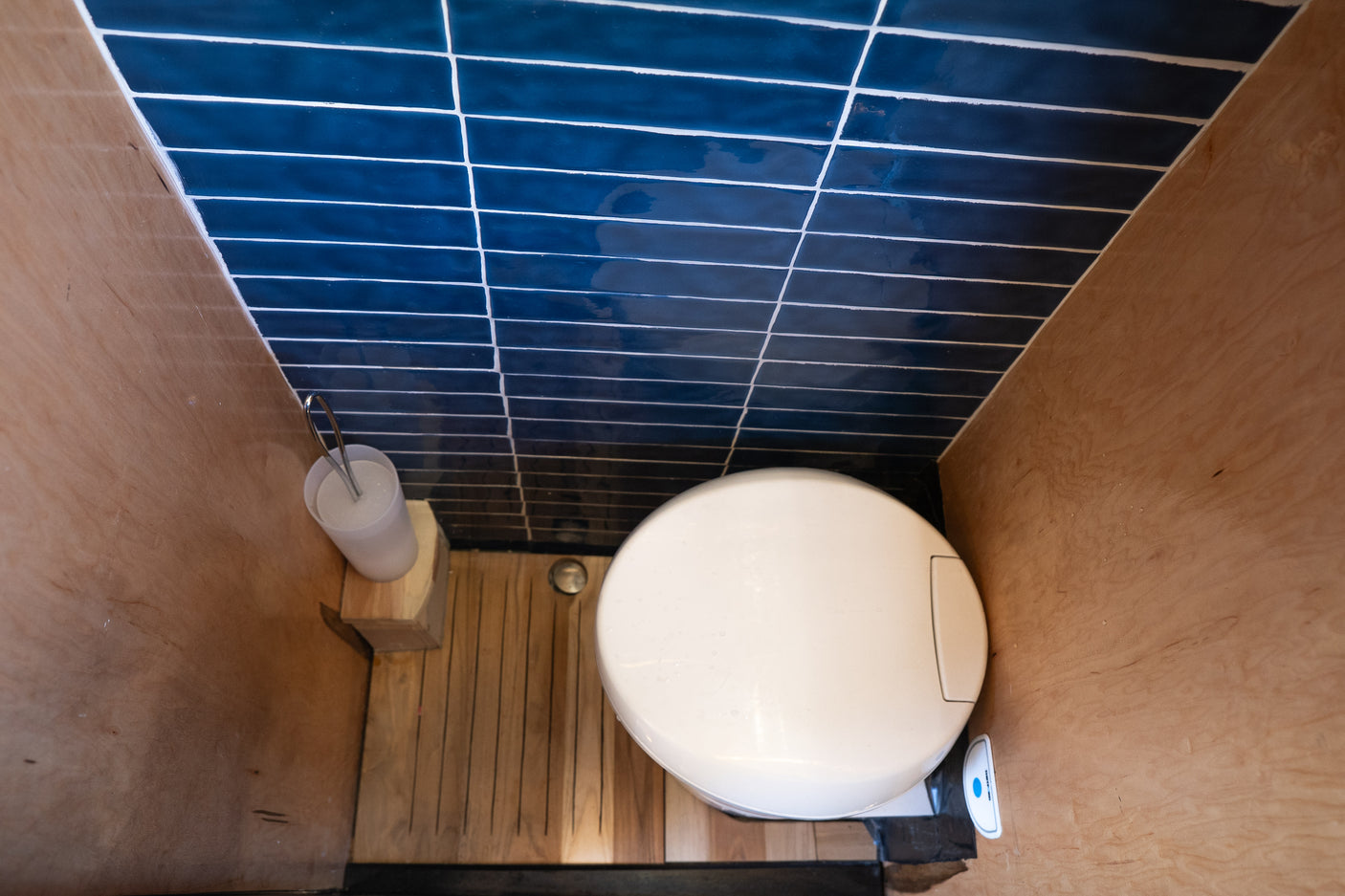 Overhead view of a compact RV rental toilet featuring a blue tiled wall, wood side panels, and a slatted wood floor.