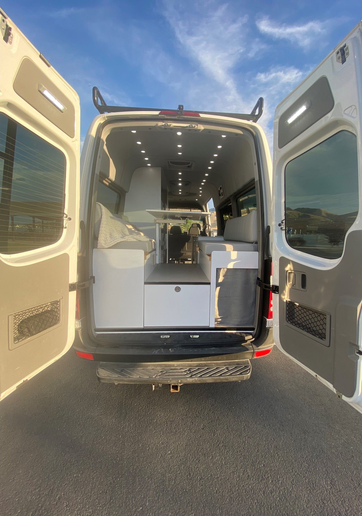 Bright white camper van interior with rear doors open, showcasing custom cabinets & abundant ceiling lights. Great for rv rental.