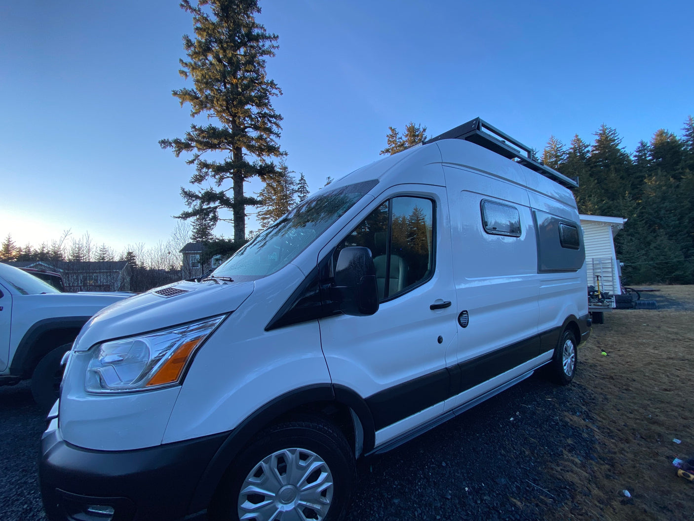 Spacious white camper van, ideal for RV rental, featuring a roof rack, parked outdoors amidst trees and blue skies.