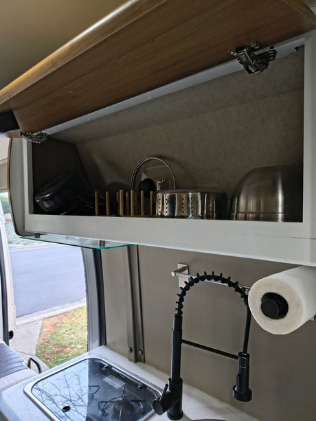 An RV rental's clever kitchen setup: stocked overhead cabinet, glass shelf, Dometic stove/sink, and pull-down faucet.