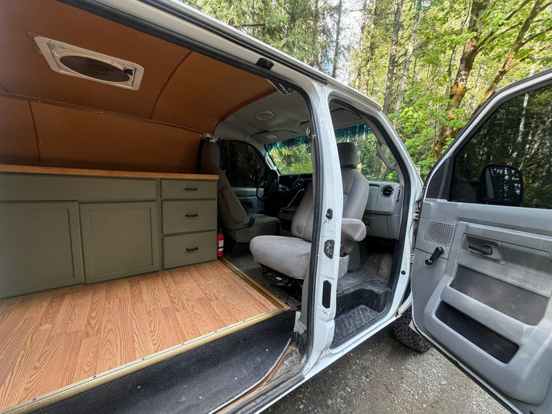 Inside a custom RV rental van conversion: wood flooring, olive cabinets, vent, and front seats, with a forest view. — photo 9