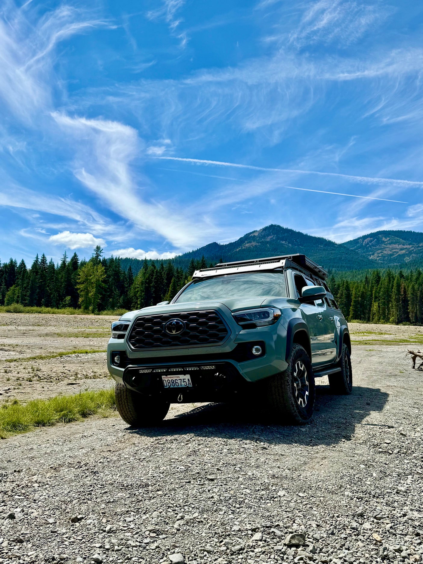 Sage green Toyota Tacoma overland truck with roof rack on gravel. Mountain and forest backdrop. Perfect for an RV rental style adventure!