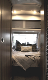 Inside an RV rental: a modern bedroom with a bed, patterned pillows, and a throw, seen from a warm wooden doorway.