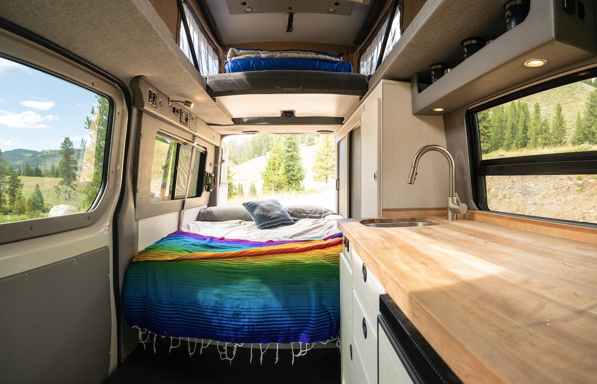 Cozy RV rental interior with a bed, small kitchen, and scenic outdoor views through wide windows, ideal for a camper van adventure.