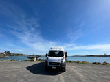 White camper van (RV rental) parked on a paved lot by a tranquil bay, featuring a bright blue sky and distant hills.