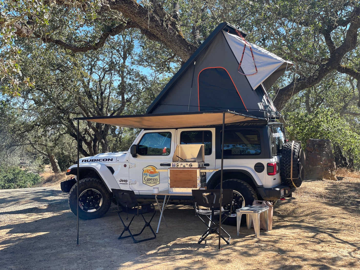 White Jeep Wrangler rv rental with rooftop tent & awning, featuring an outdoor kitchen & chairs at a wooded campsite.