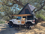 White Jeep Wrangler rv rental with rooftop tent & awning, featuring an outdoor kitchen & chairs at a wooded campsite.