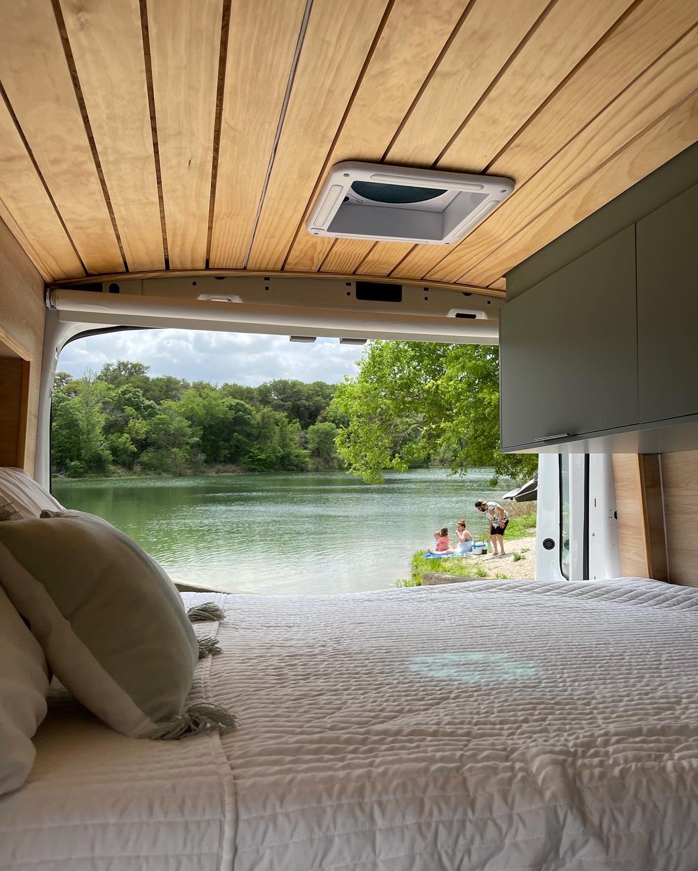 Relax by the lake! This cozy rv rental offers a stunning view from bed, with people enjoying the water's edge and lush trees.