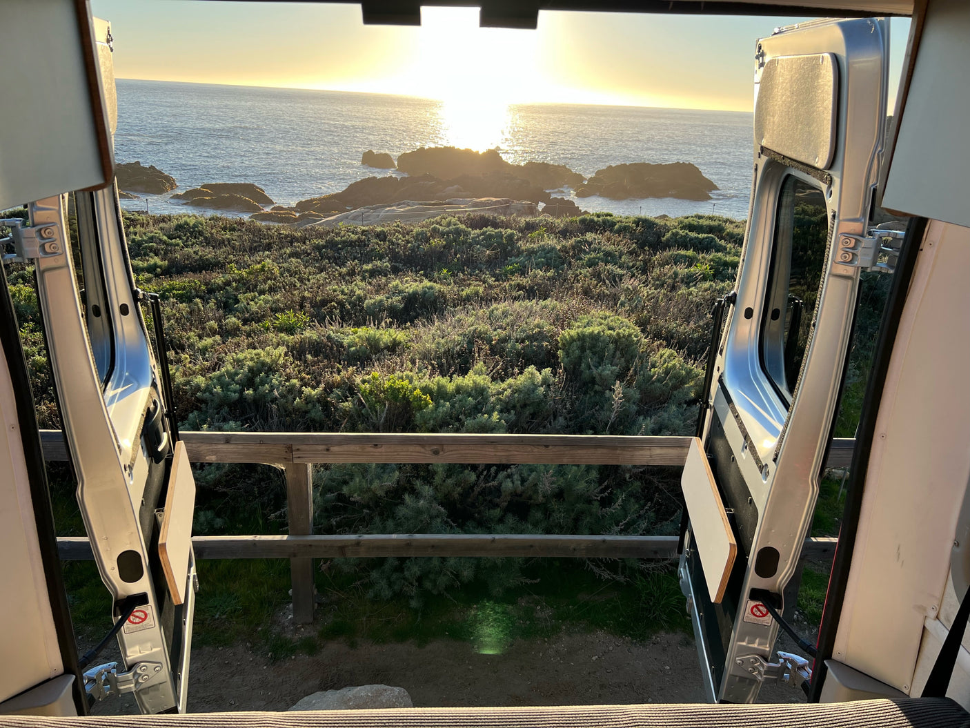 Golden sunset over the ocean, viewed from the open rear doors of an RV rental, framed by coastal greenery and rocks.