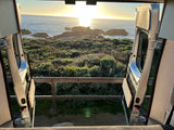 Golden sunset over the ocean, viewed from the open rear doors of an RV rental, framed by coastal greenery and rocks.