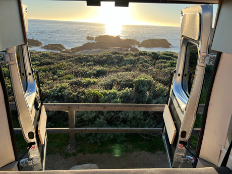 Golden sunset over the ocean, viewed from the open rear doors of an RV rental, framed by coastal greenery and rocks.