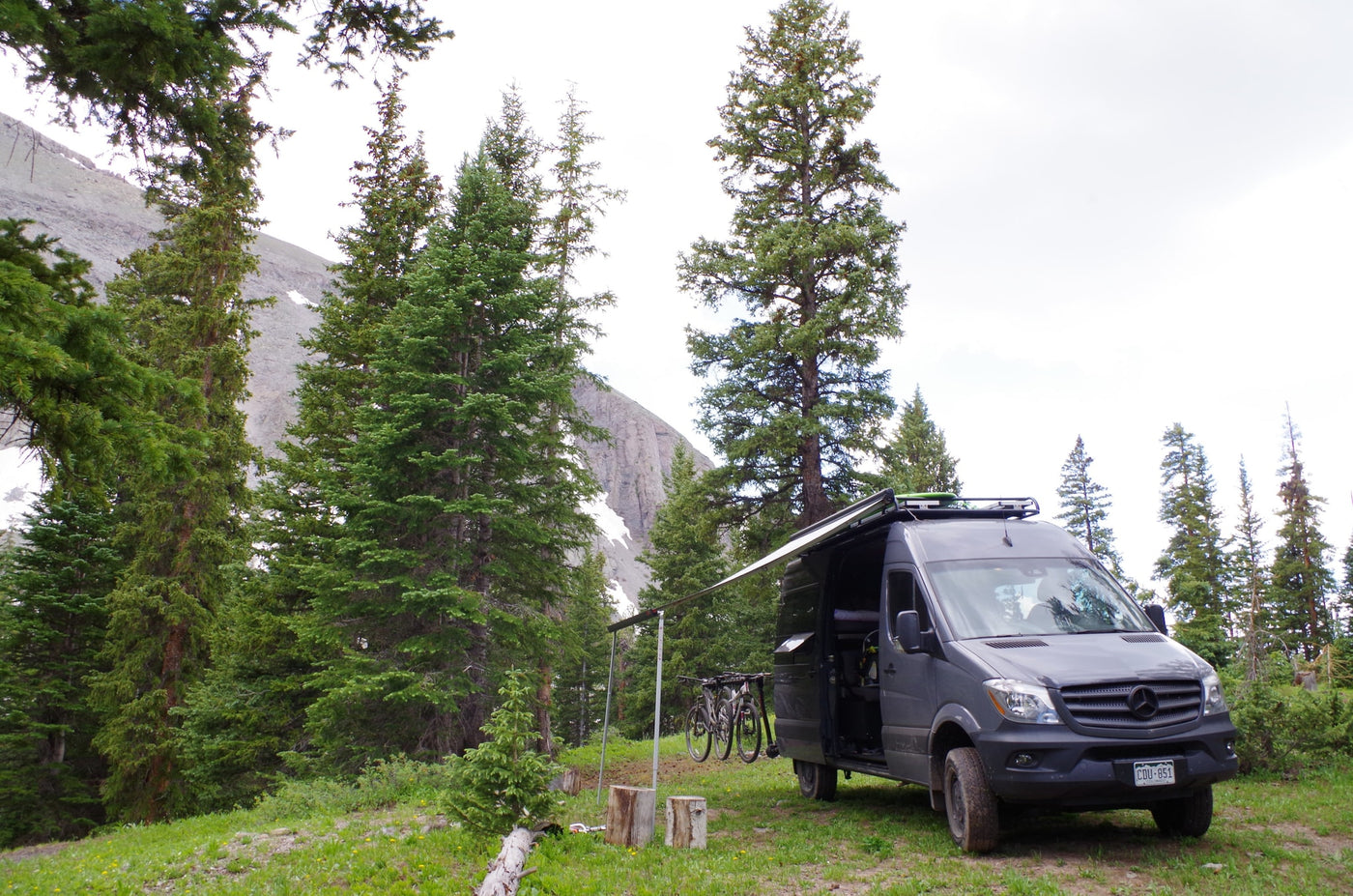 An RV rental, a grey Mercedes Sprinter van, is set up with an awning & bikes in a mountain forest campsite. — photo 10