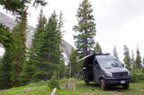 An RV rental, a grey Mercedes Sprinter van, is set up with an awning & bikes in a mountain forest campsite.