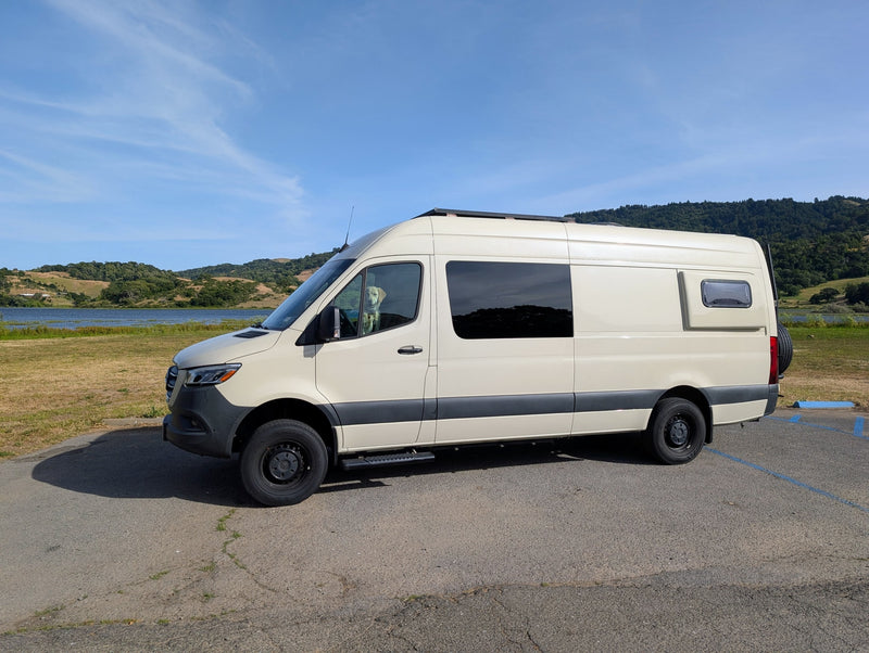 A cream-colored RV rental camper van with a golden retriever inside, parked by a scenic lake and green hills under a blue sky. — photo 8