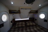 Cozy RV rental interior featuring a plaid bed, circular windows, and overhead storage for comfortable travel.
