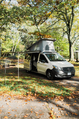 Pop-top Mercedes RV rental with awning set up at a scenic autumn campsite, ideal for outdoor trips.