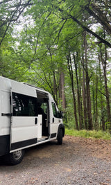 White RV rental van on a gravel forest path, side door open. Surrounded by tall green trees, ready for adventure.
