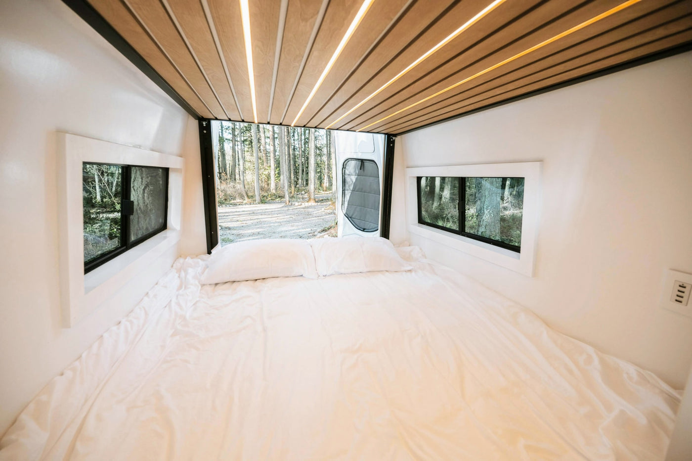 Bright RV rental camper van interior: white bed, large windows framing a forest, and modern wood-slat ceiling lights. — photo 11